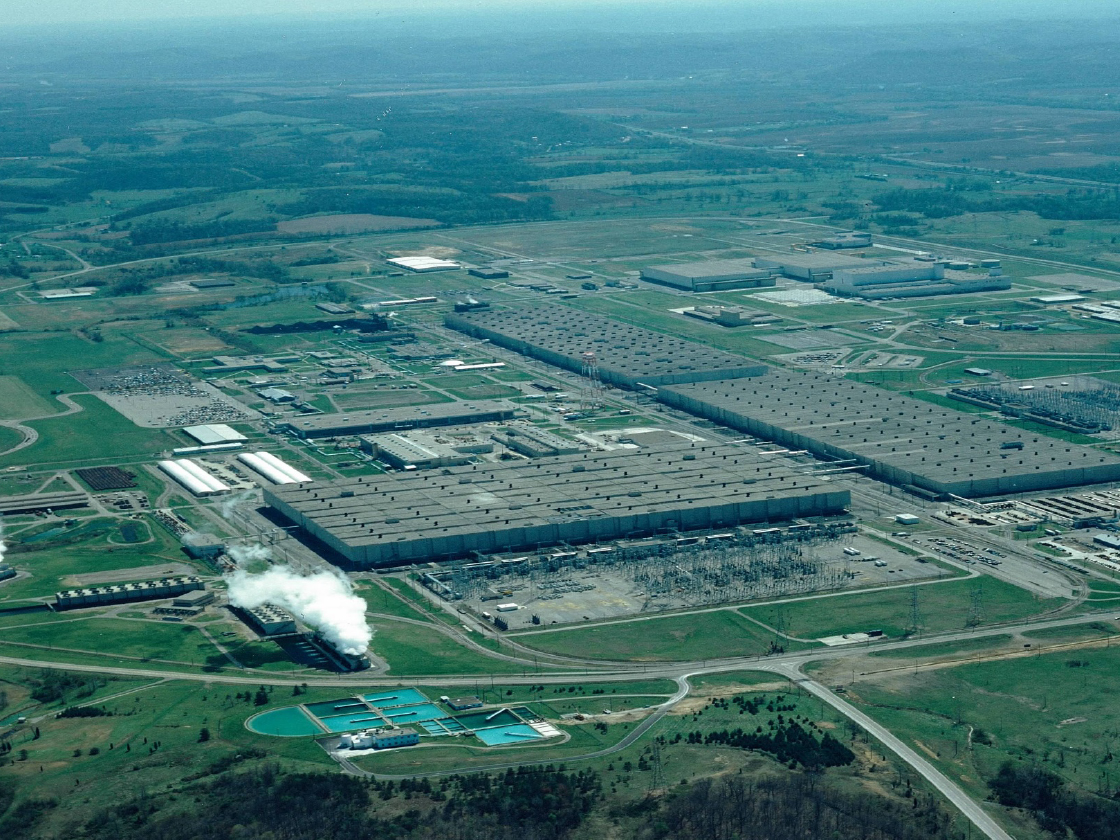 An aerial view of the Portsmouth Gaseous Diffusion Plant (PORTS) site in Piketon, Ohio, featuring several massive industrial buildings, cooling towers with steam, and the surrounding green landscape.
